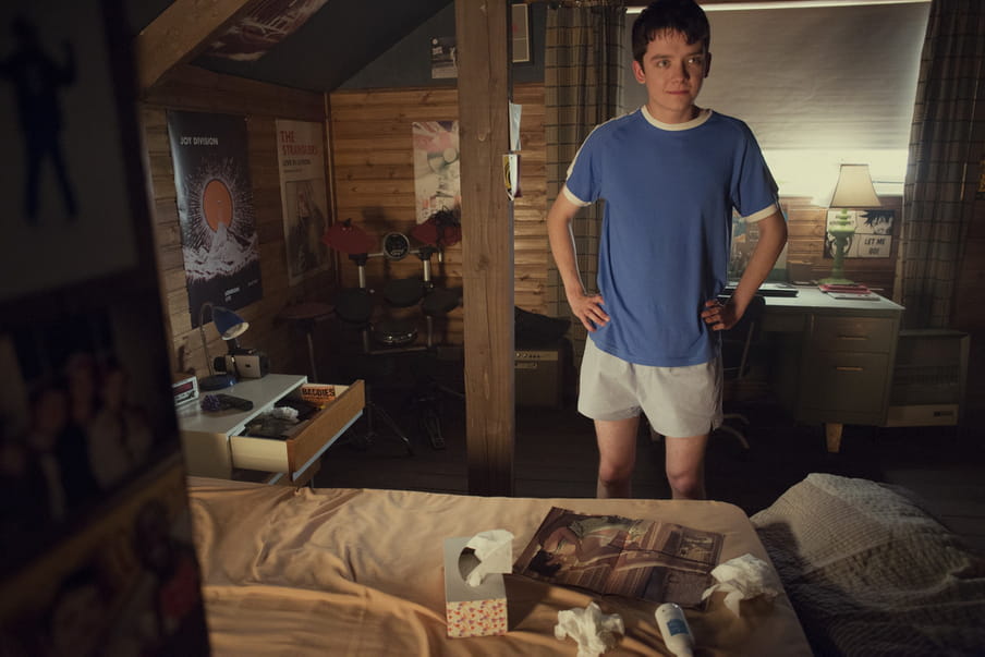 Screenshot from the series Sex Education showing a boy in his childhood room, in front of his bed. Laying on his bed are some tissues and a magazine.