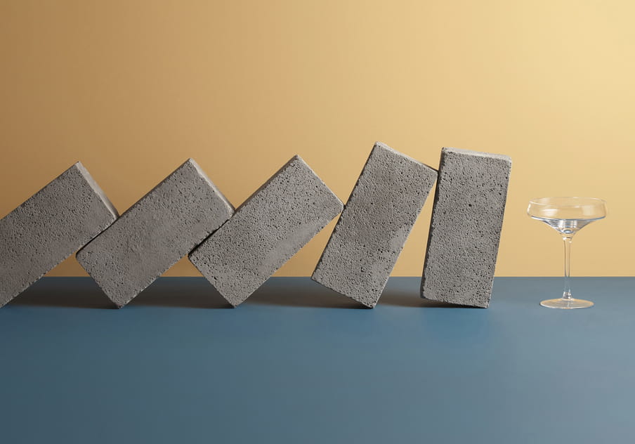 Colour photograph of a series of bricks about to fall over a champagne glass. 