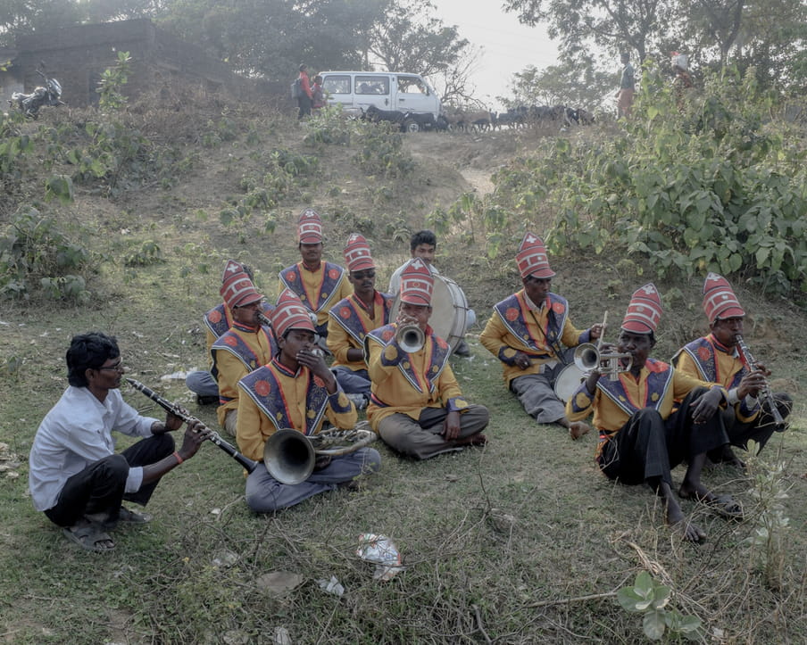 Ten Indian men sit cross-legged in a group on dry grass, dressed in a yellow, blue and pink uniform, with pink tall hats (only one plays a clarinet or flute in a simple shirt); they are part of a wind instruments ensemble, and are playing trumpets, or drums. Up the hill in the background we see a mini van.  