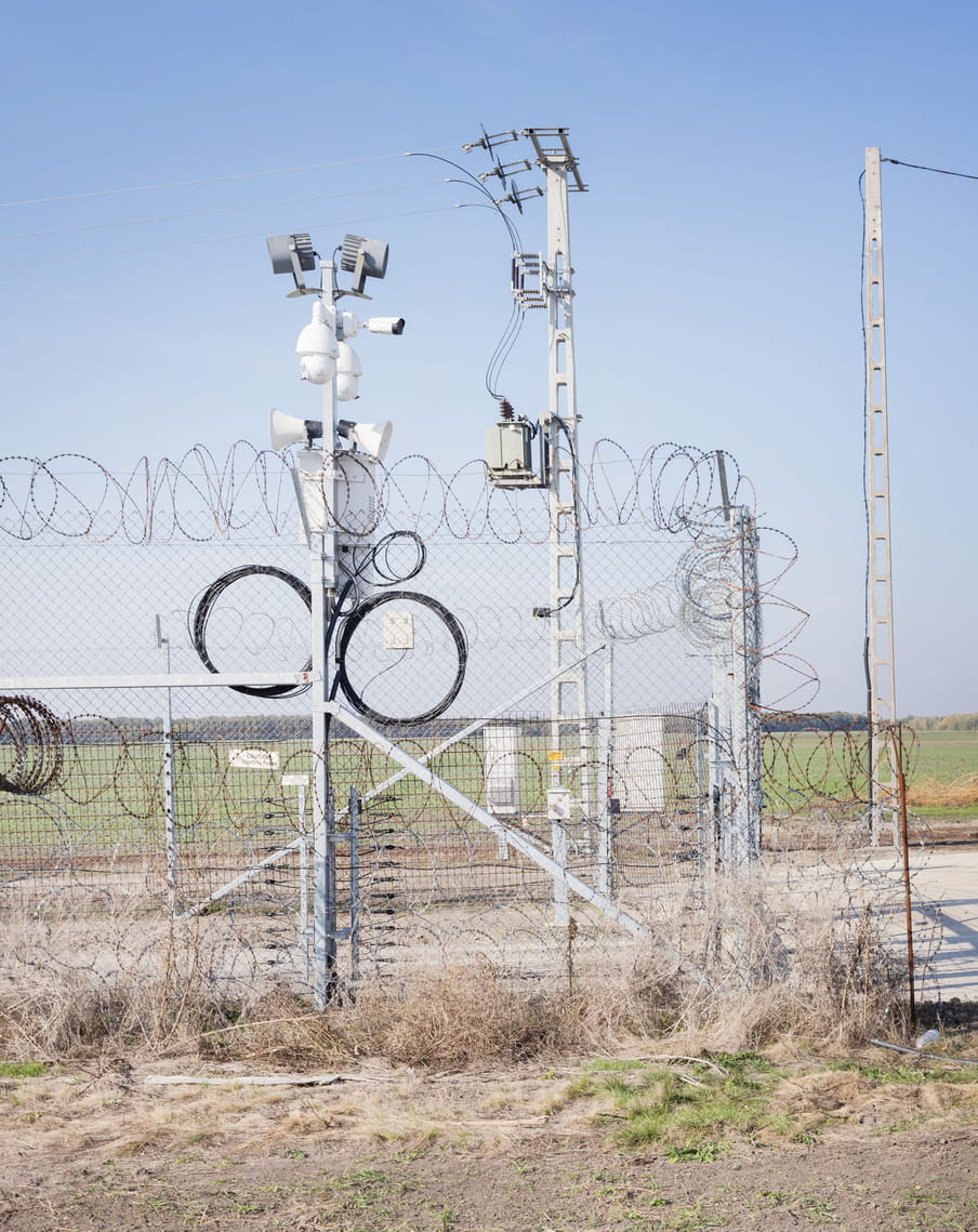Border barrier construction on the Hungarian-Serbian border. The photo shows electric barbed razor wire and masts equipped with megaphones. There is a larger electric mast in the background. Behind the construction lie a narrow grey path made of concrete and a broad, grassy field. In the background we see a thin line of forest and blue skies.