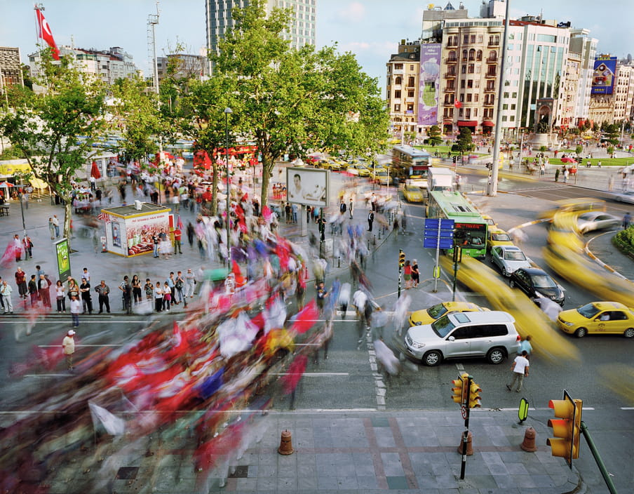 Photo of a busy street with a road trough it, yellow cabs and other cars on it. It’s shot with a high shutter speed, so people are blurred