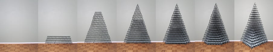 Series of seven photographs on a parquetry floor in front of a grey wall showing the progressive buildup of a pyramid made out of greyish playing cards. The first image on the left is empty and in the last one on the right, the structure is completed. 