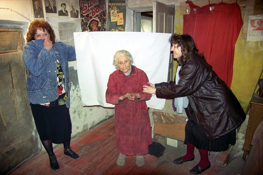 Central figure in a dressing gown, standing feebly due to her age, against a white sheet held up by two figures on either side, in a room with concrete walls and various posters on one side, and a red sheet hanging against a yellow wall on the other side