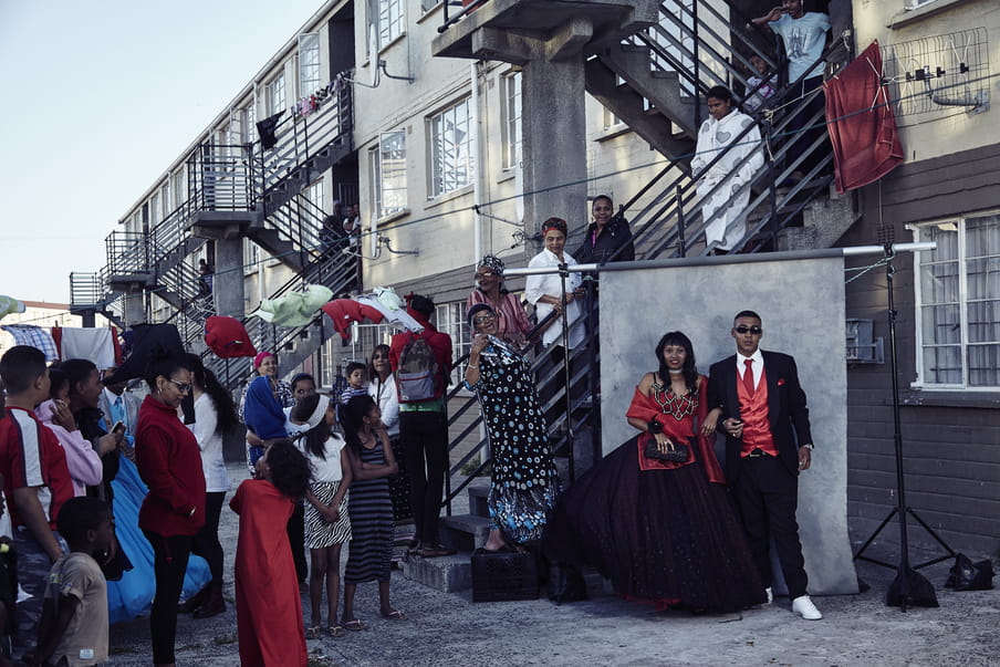 A couple all dressed up is standing in front of a grey background, ready to be photographed. People are gathered behind them.