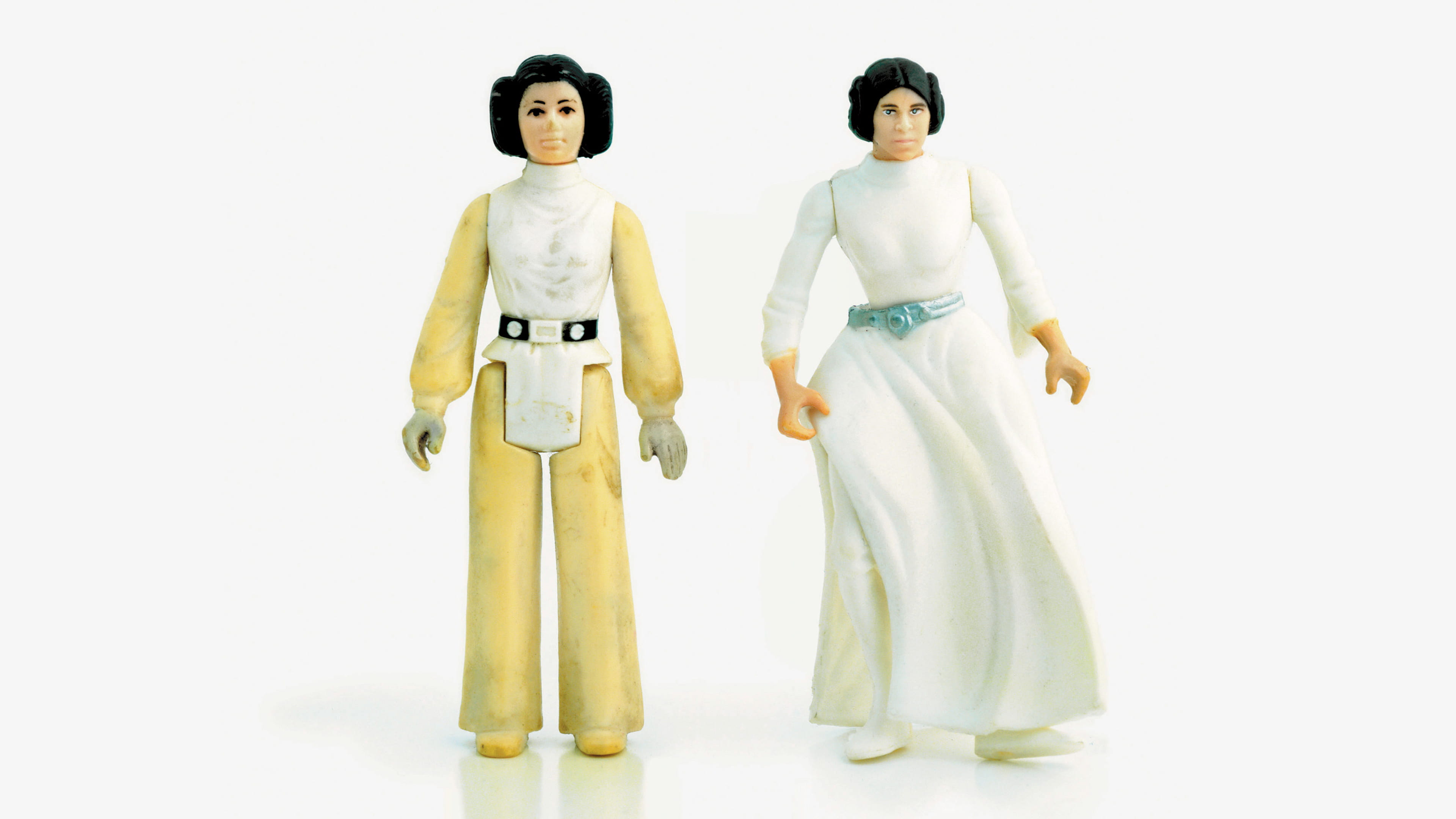 Two doll figures stand against a white background: on the left, a figure in a yellow outfit and white vest top and belt, with hair made up in buns around the ears, arms by the side. On the right, a similar looking figure, in a long-sleeved flowing white dress down to the ankles and green belt