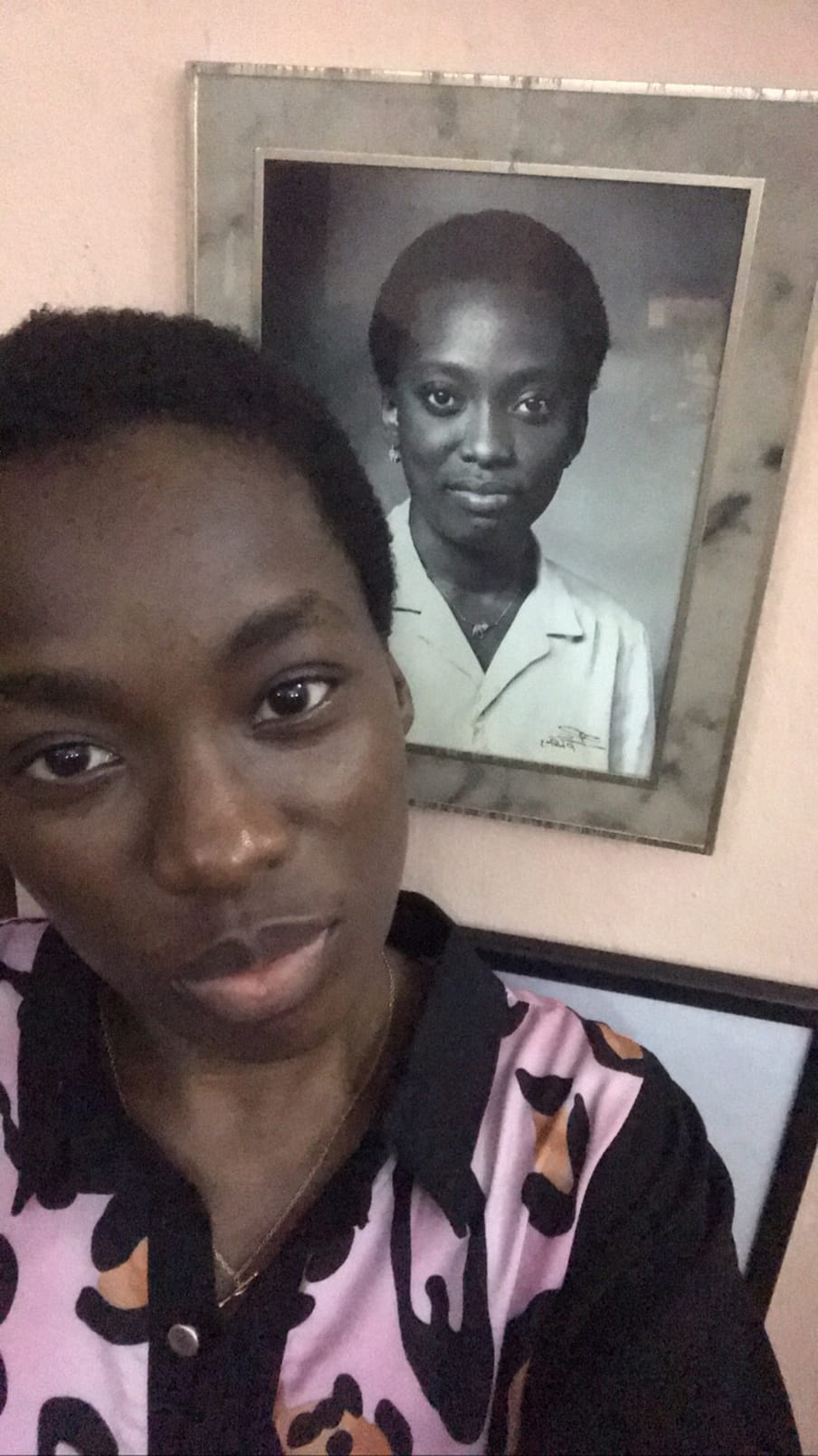OluTimehin Adegbeye stands next to a framed photo of her mother, Olusegun Adegbeye. Pictures, a woman in a black and pink top smiles at camera; behind her a black and white framed photo hangs on a wall of another woman, smiling, wearing a white shirt