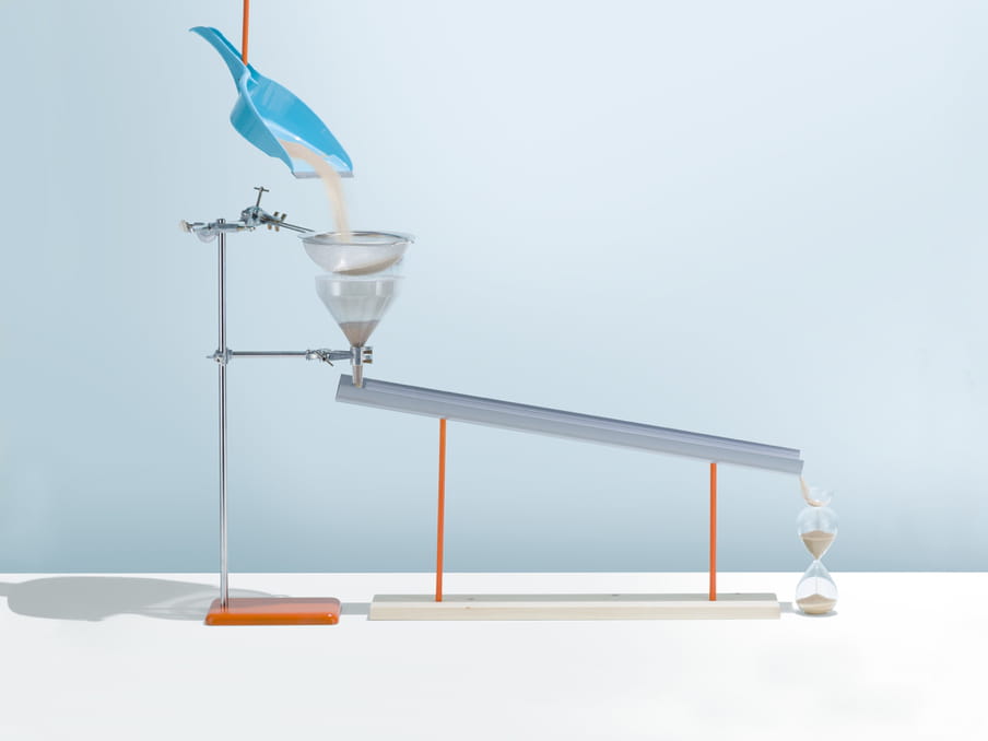 A blue dustpan is suspended from the top of this photo, a red tube pouring sand into it. The sand falls through a sieve and into a funnel, both attached to a metallic stand. The funnel links to a metal bar, pouring the sand into an hourglass on the right. The background is pastel blue, and the set-up casts shadows around.