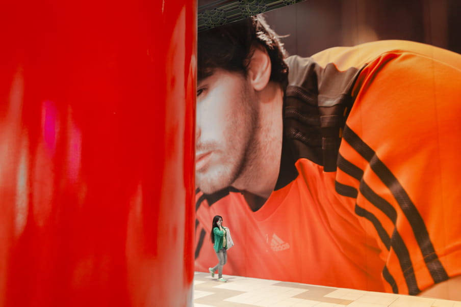 A women on the phone is walking next to a gigantic photograph of a brown-haired man wearing an Adidas orange t-shirt.