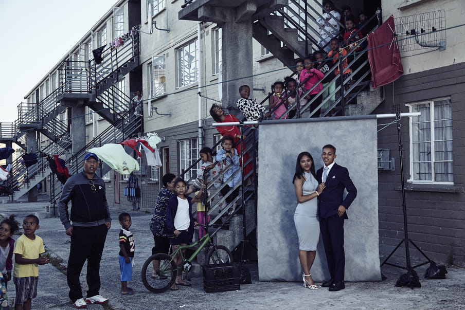 A couple all dressed up is standing in front of a grey background, ready to be photographed. People are gathered around them.