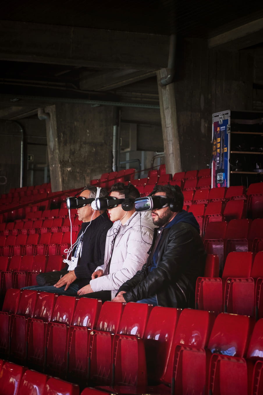 Photo of tree men sit in a football stadium with red chairs experiencing VR googles.