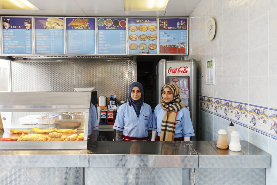 Photo of two girls wearing headscarfs, behind the counter in a fish and chips store