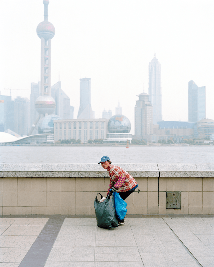 Photograph of a person slightly bent over, holding a large black plastic garbage bag. In the background there is a river and a skyline of a big city, with a tall tower creeping off the top of the screen. The sky is white