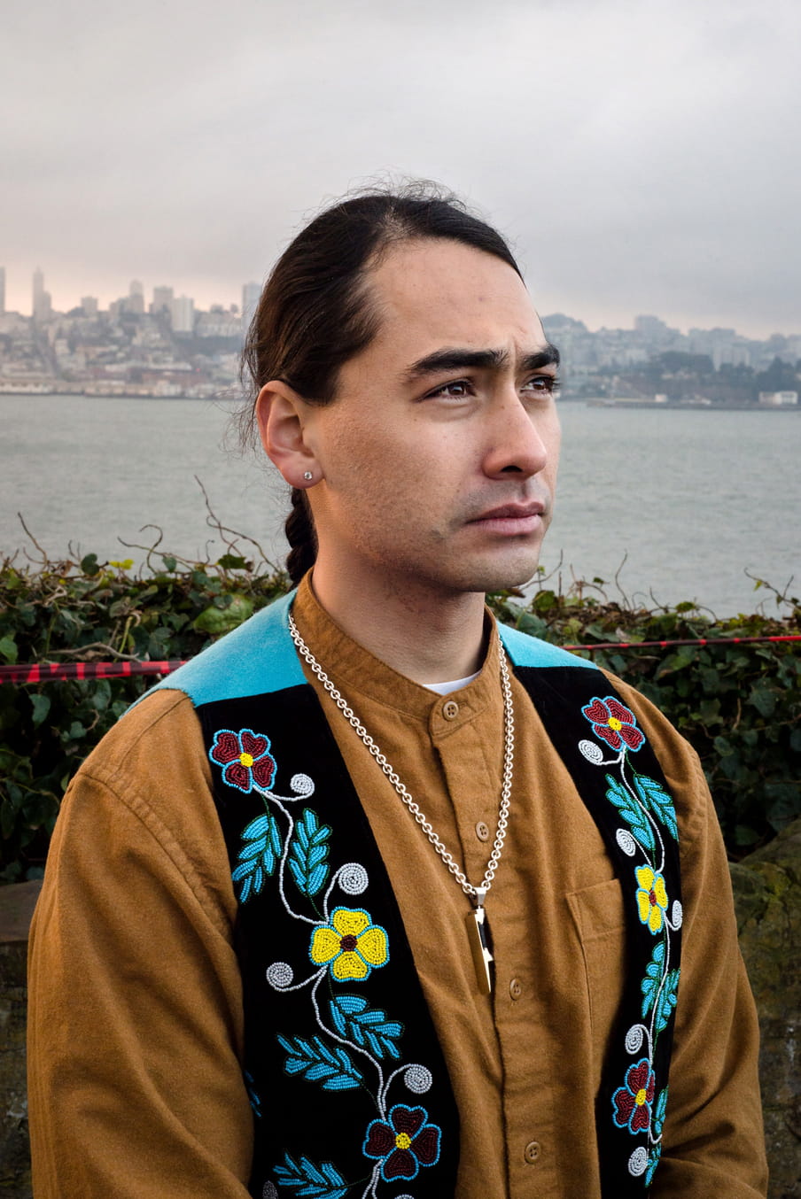 Portrait of a young man with dark long hair, a yellow shirt and a black embroidered vest - in front of water and the LA skyline