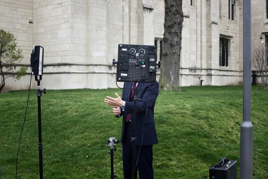 In the background you see an old looking off-white building and a tree’s bark; before it, on the grass in the centre, stands a man in a dark suit, tie and wearing a watch, hands gesticulating as if he is talking on camera. His head however is concealed behind a camera on a stand, making it look like he has a camera for a head. Around him lies various professional camera equipment such as a light, microphone, suitcase. There is a lamppost to the right of the image.