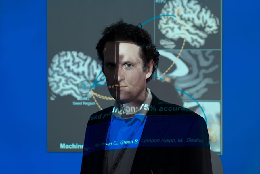 Man with short curtly hair standing in front of a beamer screen, the light of the beamer hitting his face and the screen, deforming the brain graphic on it.