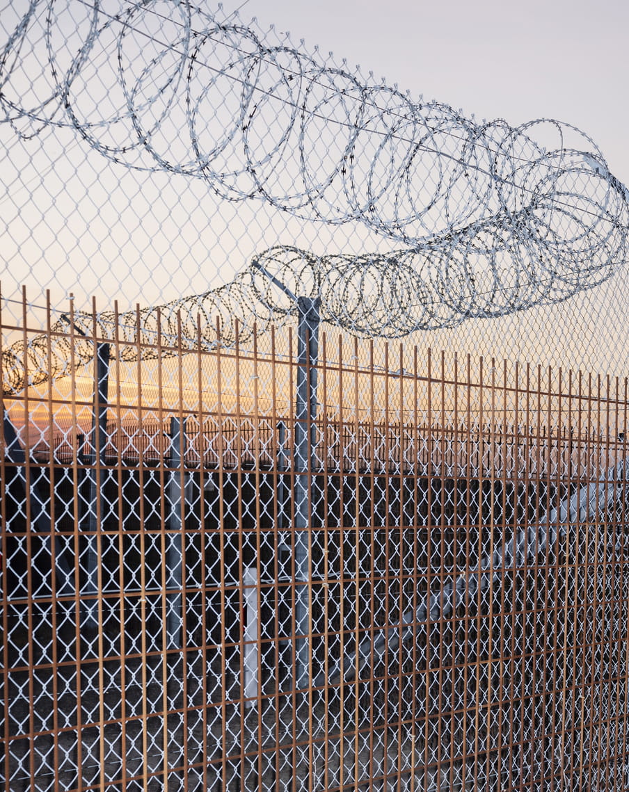 Bar a small area of light blue sky, the image is lined and crossed with different types of fencing layered across and on top of each other. There’s a brown, sturdy fence structure at the front, followed by thinner, intricate rhombus fencing that towers over the solid structure and is topped by security barbed razor wire. Thick, metallic poles uphold the barbed razor wire. Behind this claustrophobic network fencing, we see light blue sky merging into a purple orange hue from the rising sun.