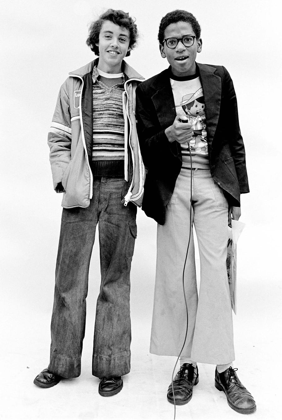Two young figures in black and white photo; to the left, in a stripy jumper, longer hair and jacket smiling at camera; the other in glasses, a cartoon shirt and black blazer, short hair, holding a camera clicker, with a surprised expression