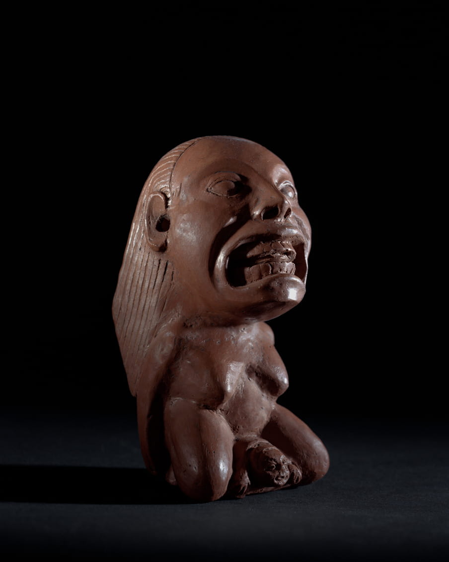 Photo of a brown statue of a female figure giving birth on her knees, her face is larger than the rest of her body and her mouth is open, showing teeth - on a black background. Compared to the last image she is sitting up more straight and the child is further in the process of being born.