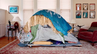 Photo of a living room of a middle or upper class house, with homeless shelter in it made up from different pieces of cloth and an old tent