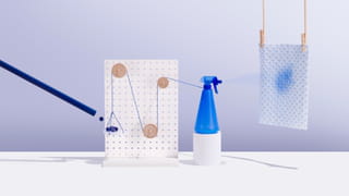 Photo of a blue tube on the left, throwing a small blue ball onto a white, round platform held in place by a blue string. The blue string is whirled around three wooden circular pegs, handing in various places on a vertical, rectangular white wooden board with holes in it. The string is connected on the right to the nozzle of a blue spray bottle. The blue and white kitchen checked kitchen towel hanging to the right of the picture has a blue spray stain on it.