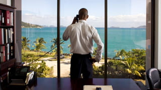 Photo of a man standing in an office in front of a window overlooking a bay with palm trees
