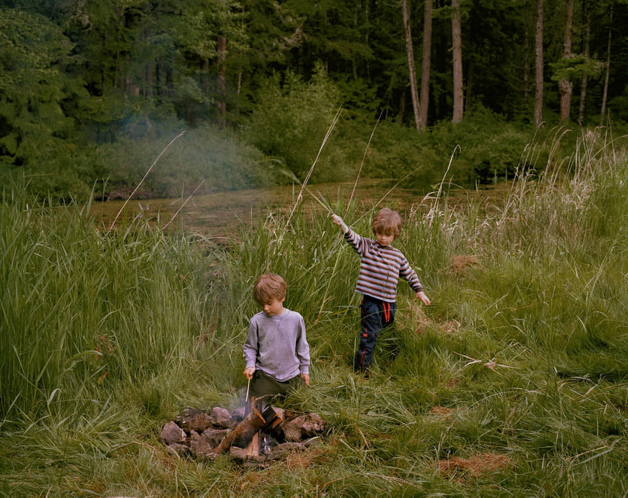 Photo of two boys playing in a field, one is poking a fire with a stick