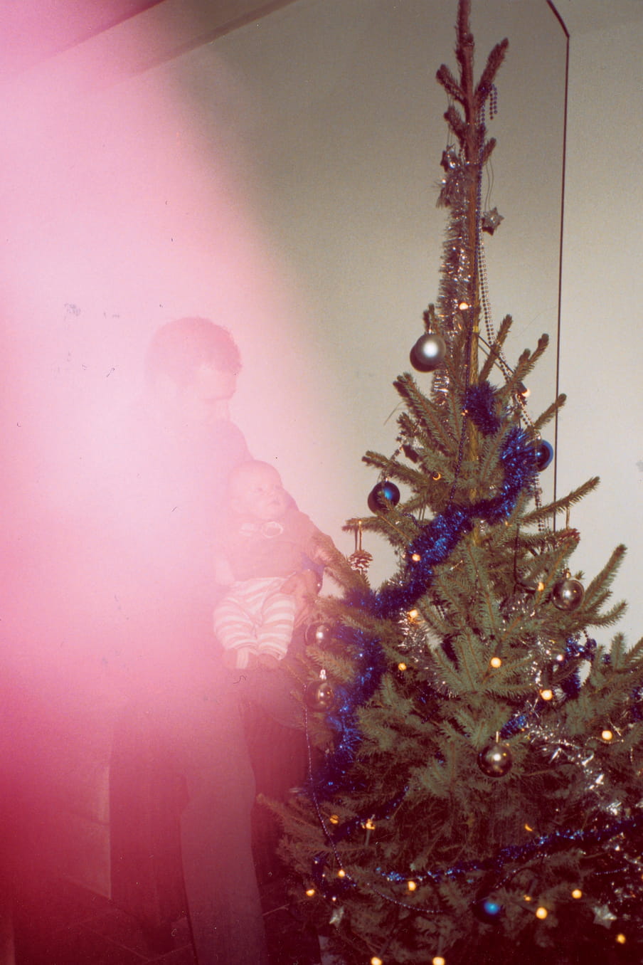 Photograph of a man holding a child next to a christmas three, a red light flare blocking the view of the man with child.