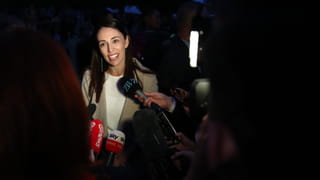 Photo of a woman with dark hair and red lipstick, reporters around her holding microphones up to her face.