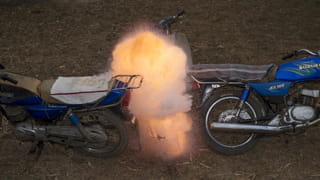 The ground is grassy with brown straw, upon which we see two blue Suzuki motorbikes, both not fully in shot. Where they touch, there is a fire which looks like it has just combusted