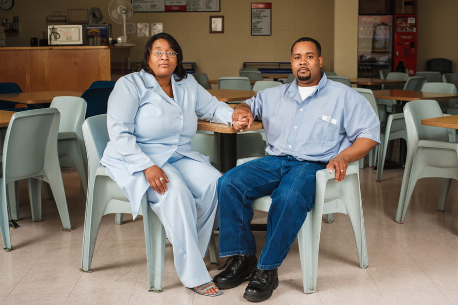 Photo of a man and a woman sitting on plastic chairs, holding hands across a table. The woman is wearing a light blue suit. The man is wearing his prison uniform, jeans and a blue shirt. They are sitting close together.