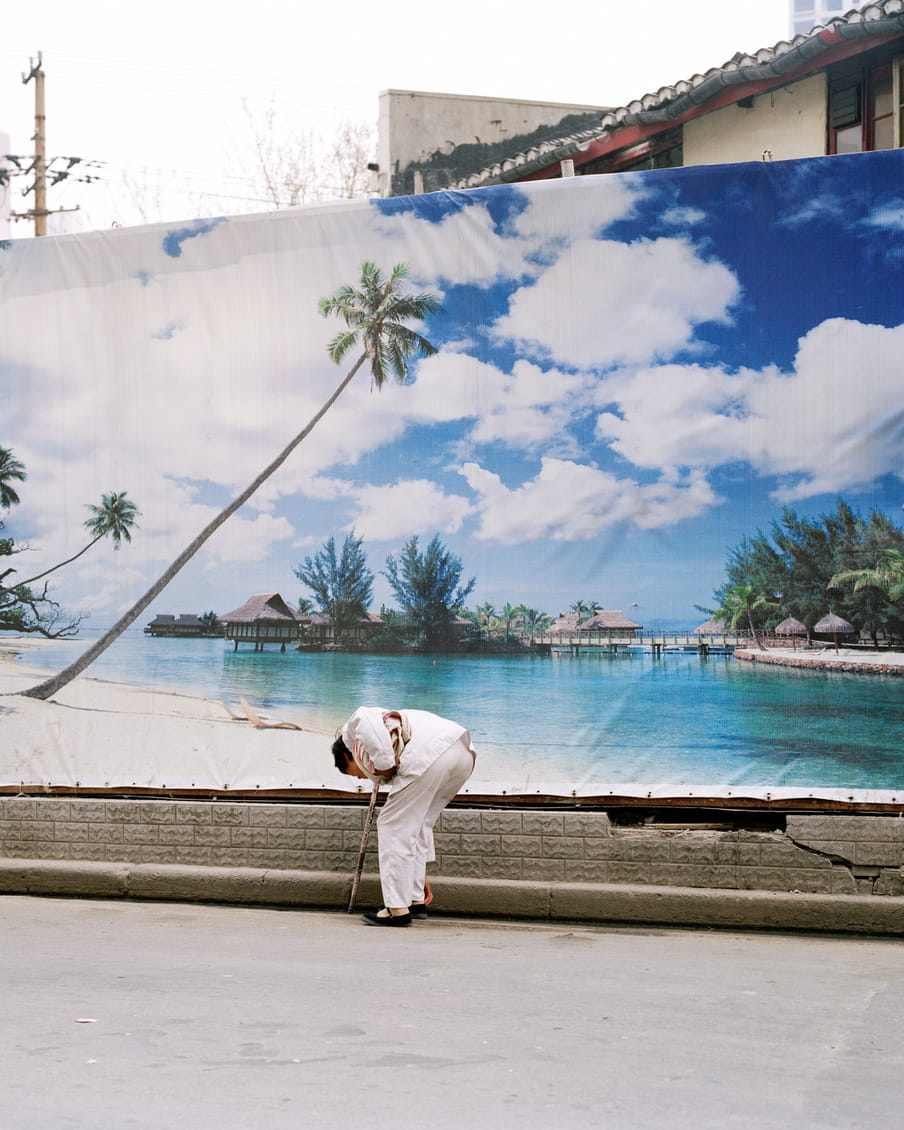 Photograph of a person picking up something from the road. In the background there is a large banner that depicts a tropical beach with small houses, part of a roof peeks out above the banner.