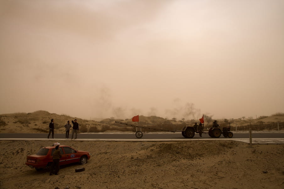 A truck and a car are park on a road in the desert. A few people are standing by. 