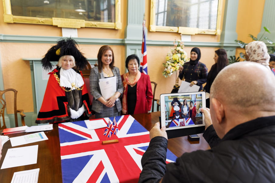 Photo of a woman being photographed in front of the UK flag, holding a paper. 