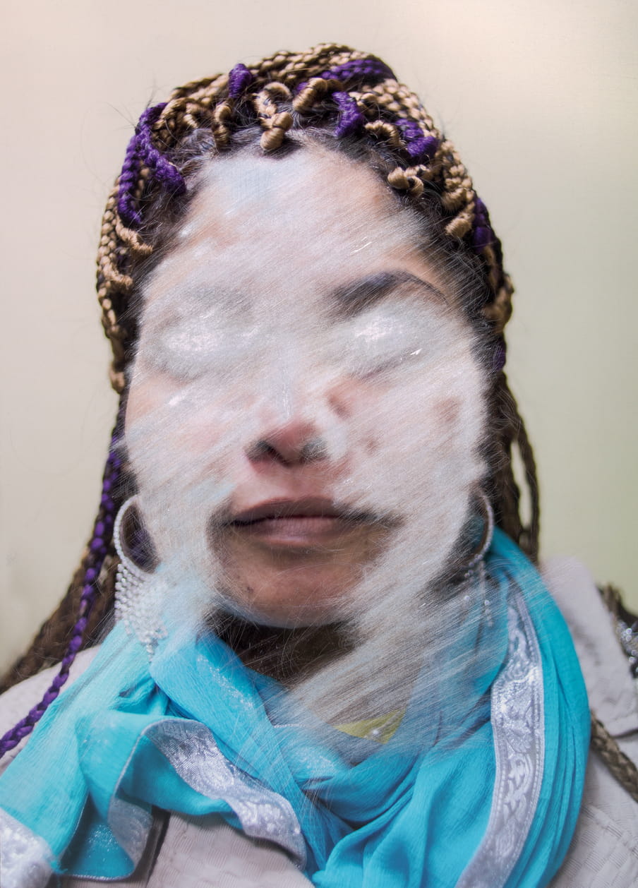Colour portrait of a woman with purple and blonde dreadlocks, silver earrings and a blue scarf, in which her face is unrecognisable because of white scratches on it, leaving only a faint smile exposed.