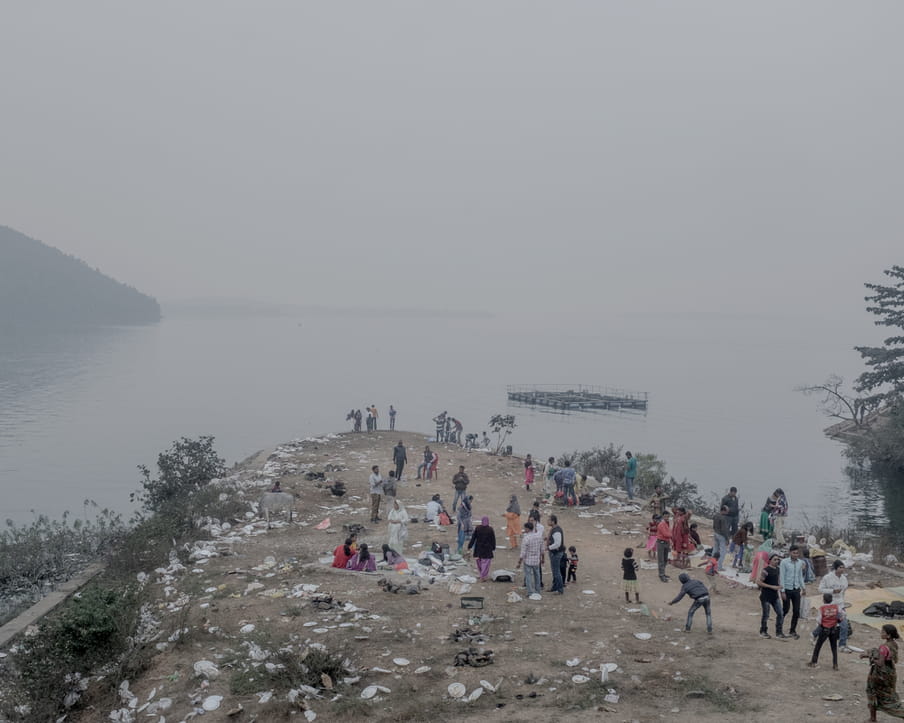 Colour photograph of a group of people near the water. Some are standing while other are sitting on the group on blankets, others are arriving or living. The grond is covered by plastic plates and other white trash. In the background, a platform is floating on the water.