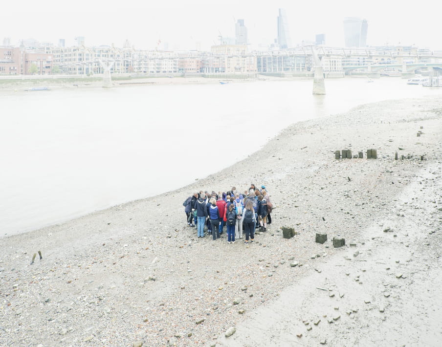 Group of people standing close together on a beach with pebbles, the skyline of a city showing