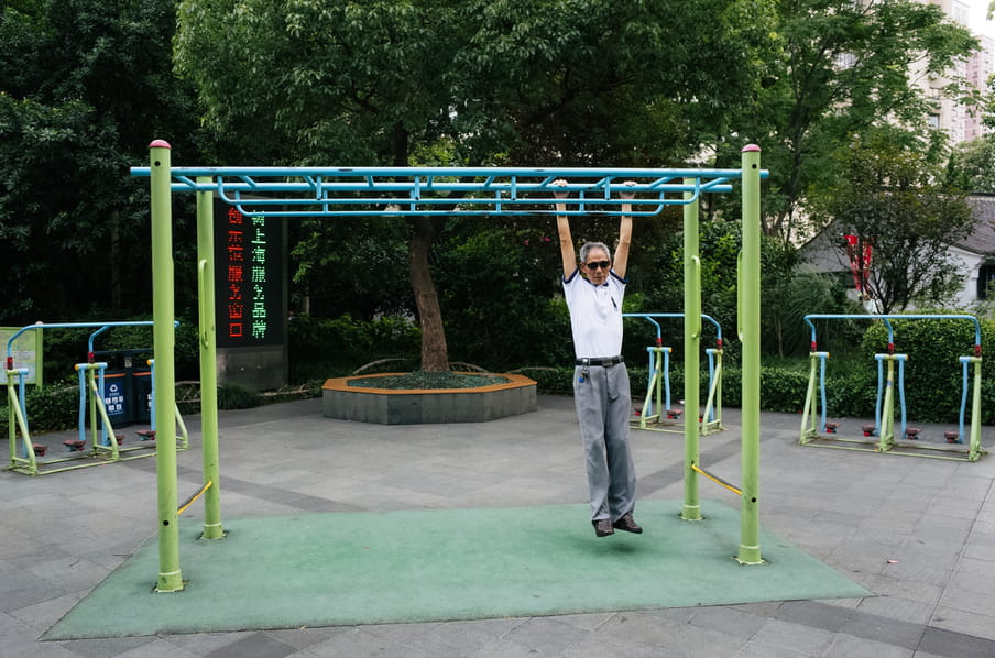 An old man in an outdoor exercise park, swinging from a high bar