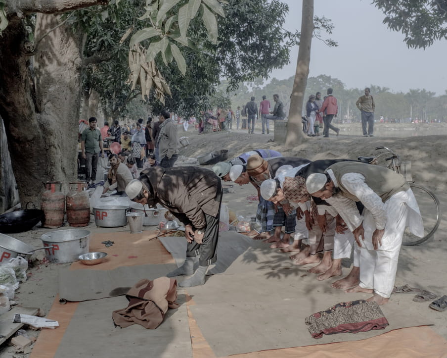 Colour photograph of a group of men bending forward in a praying position. They all wear a white Taqiyah and stand on beige mats. Next to them are gaz tanks and big metallic pots. Trees and passerby are visible in the background. 