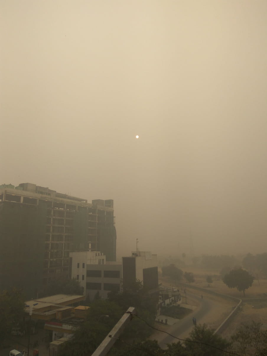 A picture showing the toxic smog in Delhi, shot on a morning in November 2016. The shot is taken from the balcony of a house on the sixth floor of a residential building. It is shrouded in a thick greyish-yellow tint that covers the building, trees, and people in front. The sun is a tiny, dim blob in the sky that looks more like the moon.