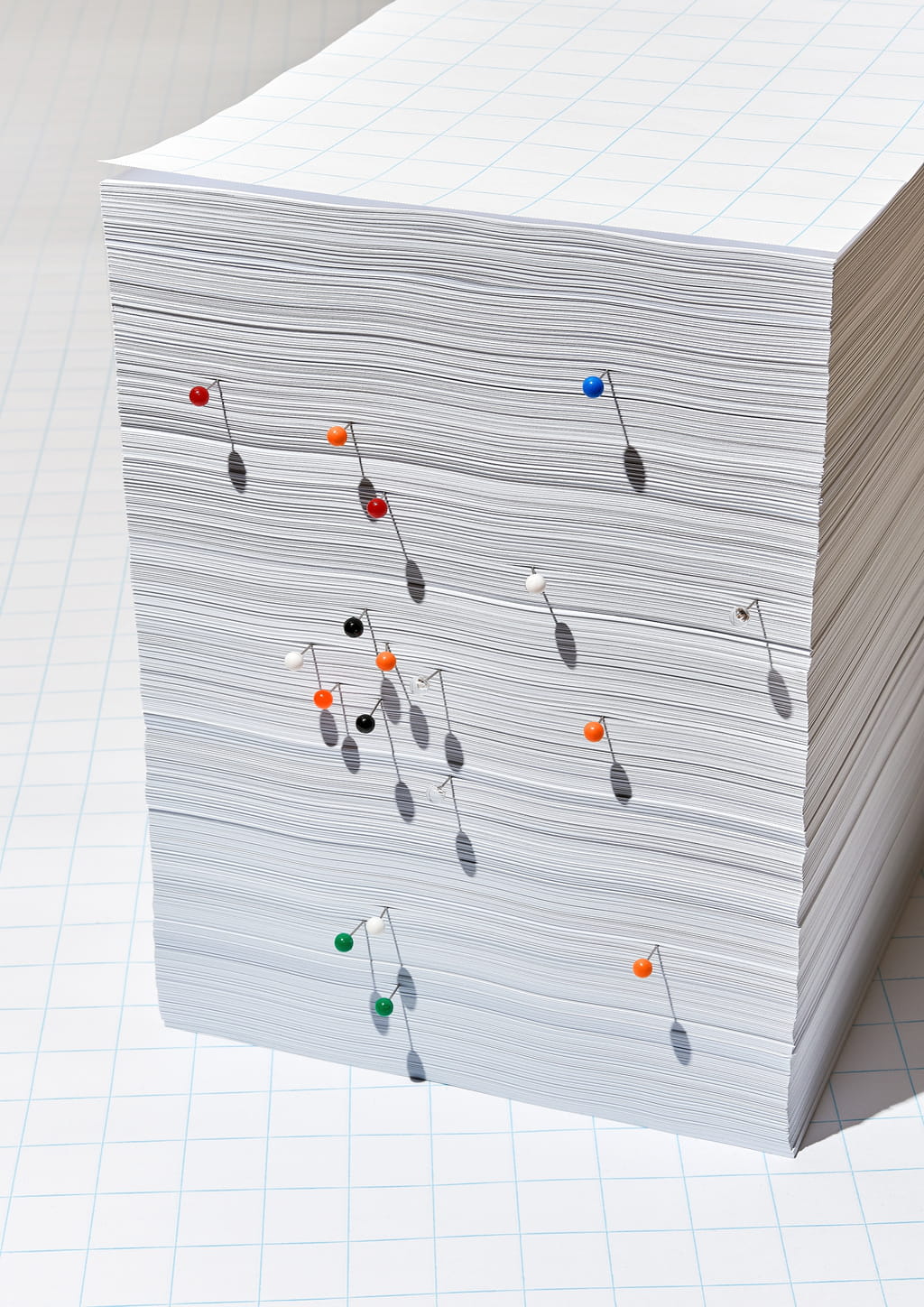 Photograph of a stack of gridded white paper with colourful needles inserted in it.