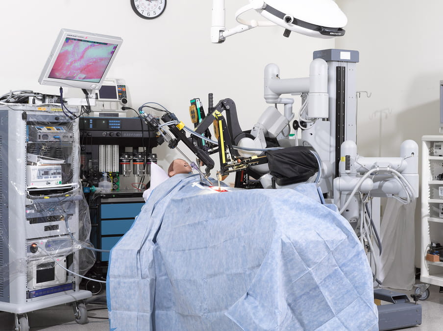 Photo of a robot doing an operation on the bloody ‘stomach’ of a mannequin, which is covered in a blue paper cloth and surrounded by machines, cables and hospital equipment, including a clock on the wall and a monitor above
