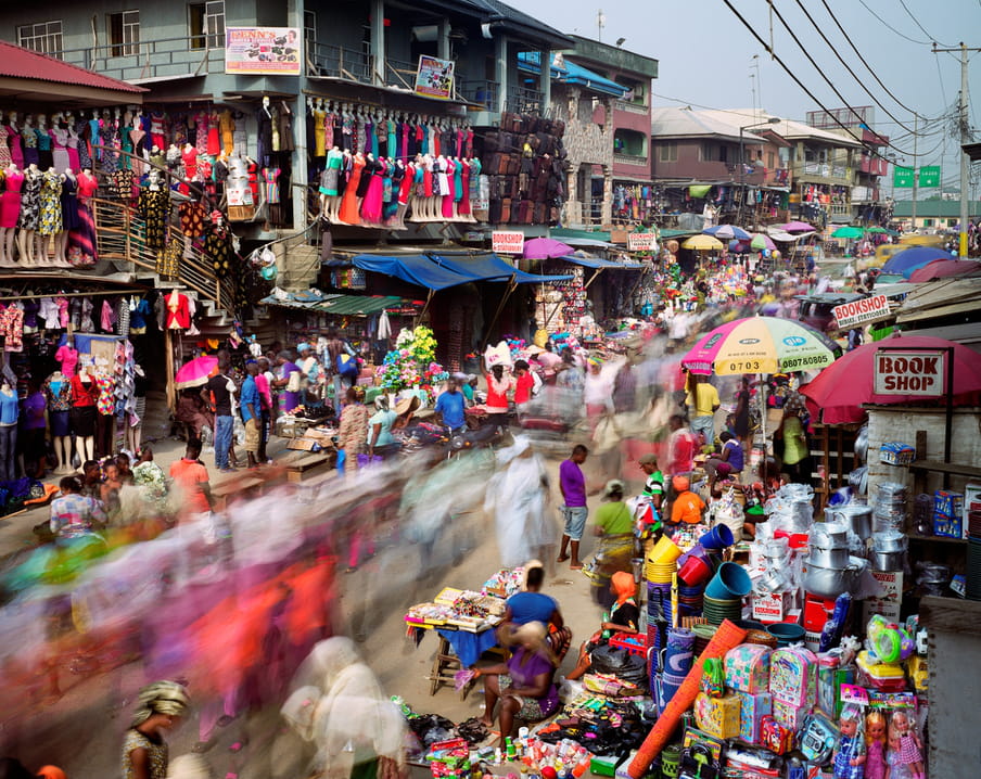 Photo of a busy street with a market on it, colourful merchandise hanging everywhere. It’s shot with a high shutterspeed, so people are blurred