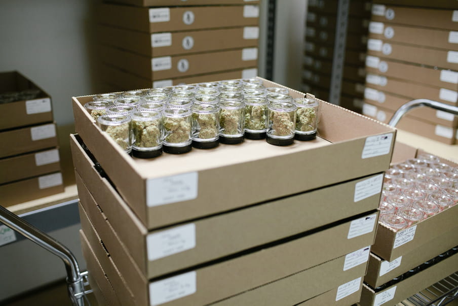 Photo of paperboard boxes stacked on each other, some boxes filled with plastic containers with cannabis, some are filled with empty containers.