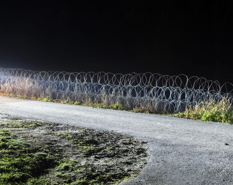 The photo highlights an area on the Hungarian border. We see a small, grey, concrete road branching to the left. In the belly of its curve lies a muddy grass patch. It’s night, but there’s artificial light making the area visible. We don’t see the source of the light. On the right side of the road, we see three rows of round, barbed razor wire stacked on top of each other against pitch black night sky.