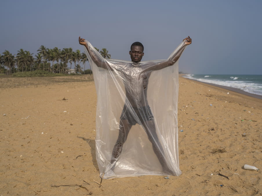 Figure of a person standing in a star shape on a sandy beach with the ocean and trees behind him, wearing shorts, and he holds a big sheet of transparent thin plastic film against his body; only his head and tops his hands are uncovered, and he has his eyes closed