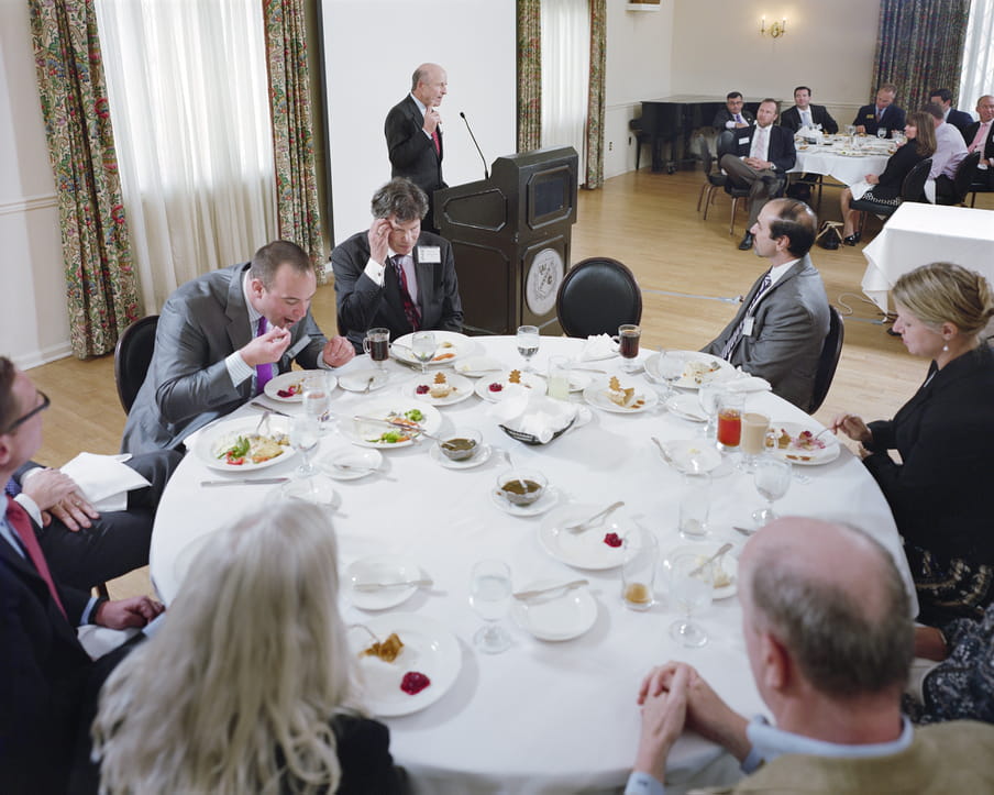 Picture of a round table with a white table cloth, men in suits and women in professional clothing around it. A man is speaking to the crowd.