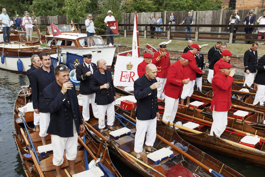 Photo of a group of men on a boat, taking a sip from their glass. Half of them are wearing a red and white suit, the other half black and white.