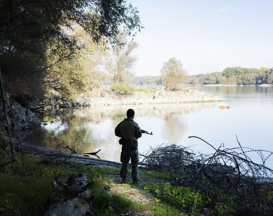 Image of a Hungarian Special Forces soldier in full uniform holding a rifle. We see his back as he’s stood alone on a patch of sandy earth at a lakeside, an area on the Croatian-Hungarian border that he’s patrolling. He’s looking out at the calm lake that’s framed by thick, leafy forest. It’s a sunny day and the image shows light blue sky. The man is surrounded by some grass and wood and there are clusters of barbed razor wire lining the ground on his left.