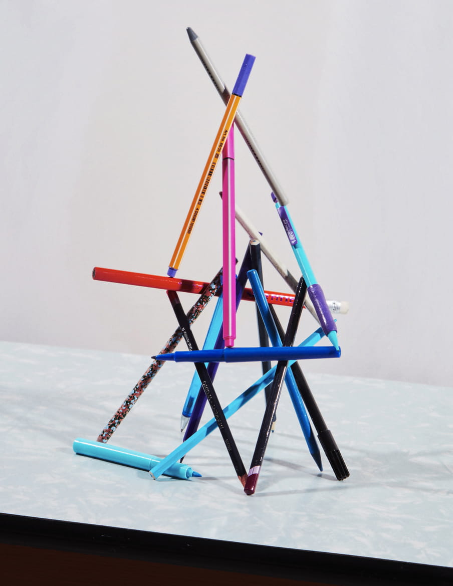 Colourful pens and pencils are stack together, in balance, creating a pyramid. The structure lays on a light blue and white marble looking like surface. 