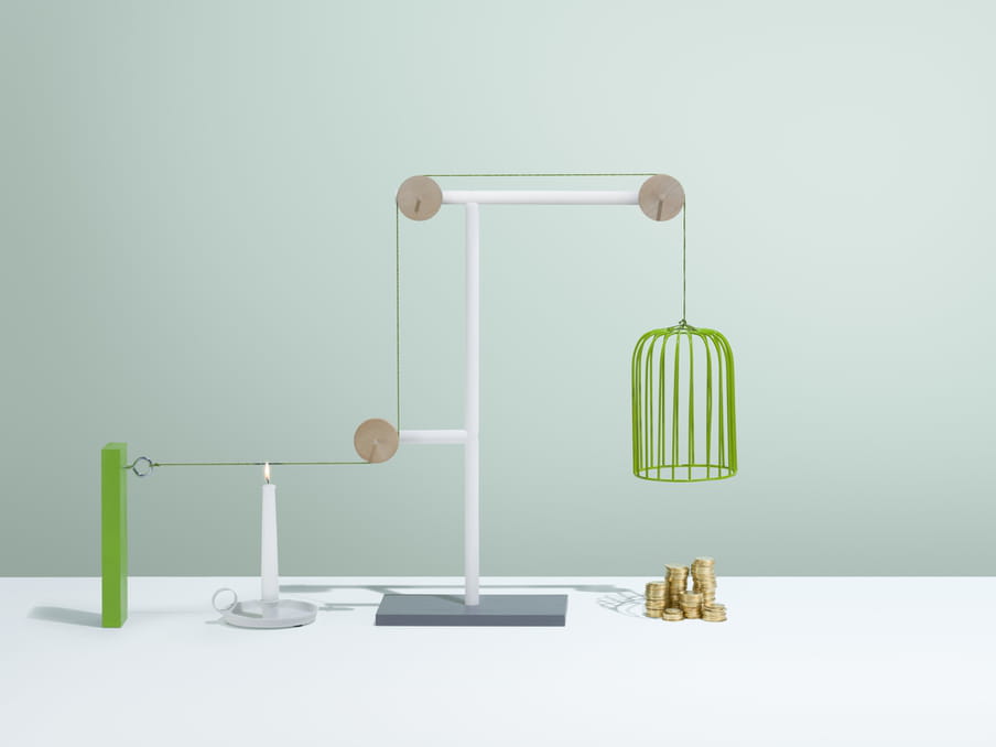 An object looking like a long lampshade made of green wire awaits above five piles of gold coins of different sizes. The lampshade is held in place by green string suspended over three wooden circular pegs connected at various lengths on a white stand. A lit candle stands to the right below the green string, ready to burn the string and drop the cage like object onto the coins.